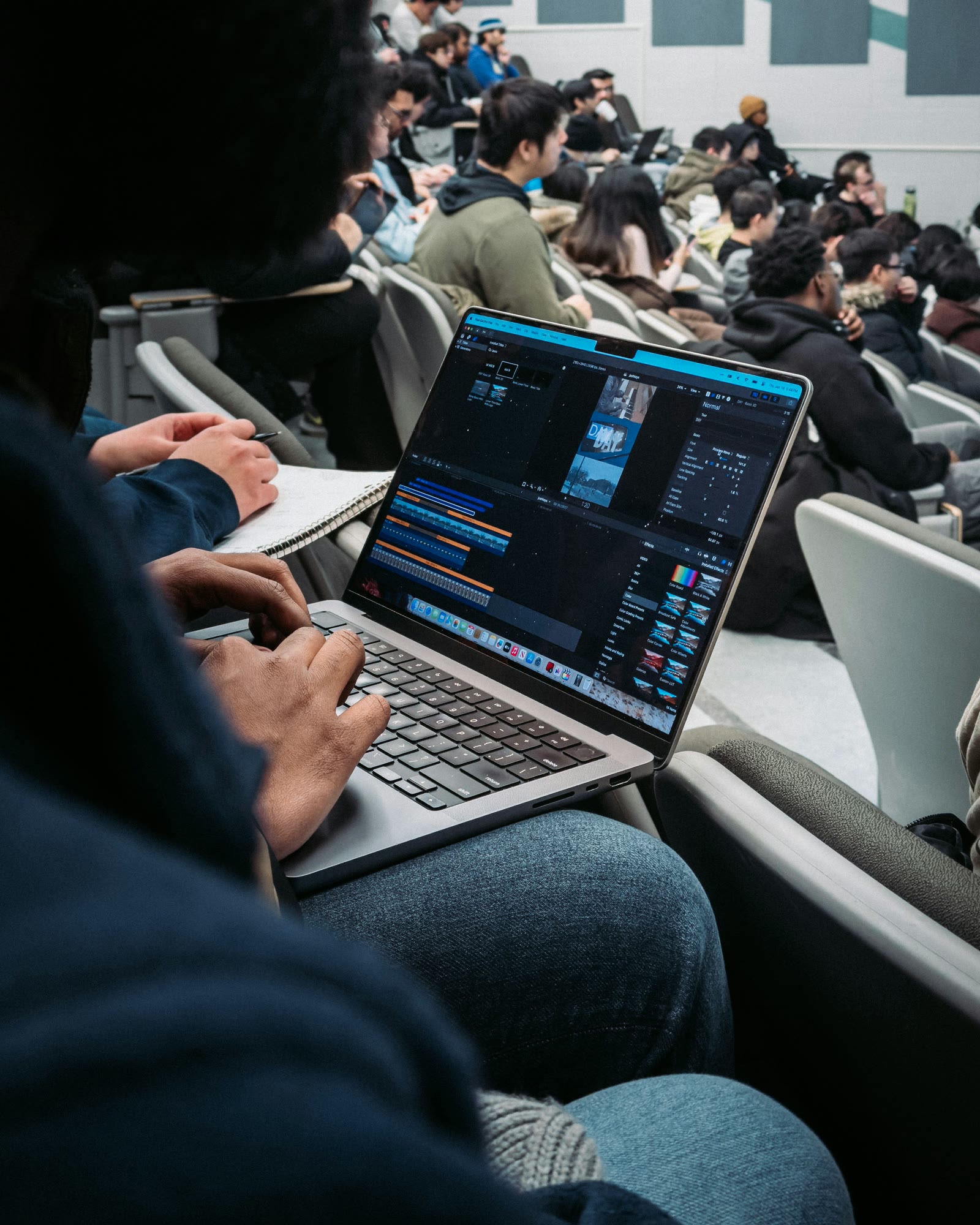 A professional editing on a laptop in a lecture hall, the Sierra pin on their jacket sleeve.